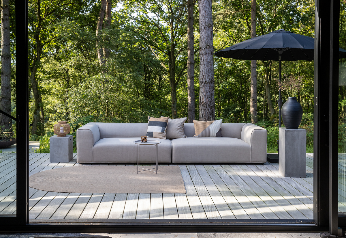 Lys grå modulsofa på træterrasse med puder, lille sidebord, rundt tæppe, sort parasol og stor vase; skov i baggrunden.