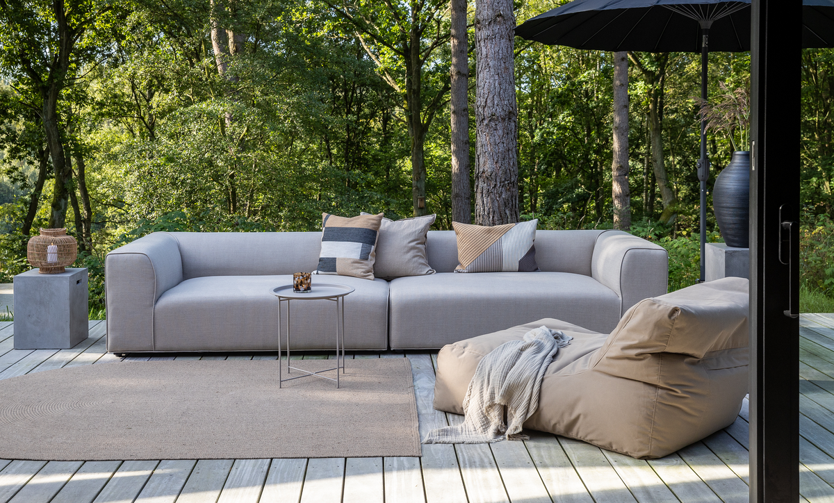 Lysgrå modulsofa på træterrasse med puder, rundt sidebord med lys, beige sækkestol med plaid og sorte parasoller i grøn skovomgivelse.