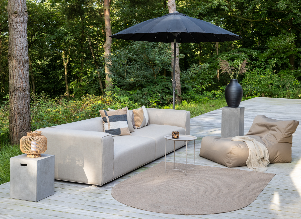 Udendørs terrasse med beige sofa og sækkestol på trædæk, sort parasol, rundt sidebord med lys, neutral måtte og sort vase med pampasgræs foran grøn skov.