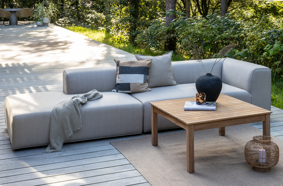 Udendørs lounge på træterrasse med lysegrå modulsofa, teak sofabord, puder og plaid, sort vase med strå, bog og lyslygte, grøn beplantning i baggrunden.