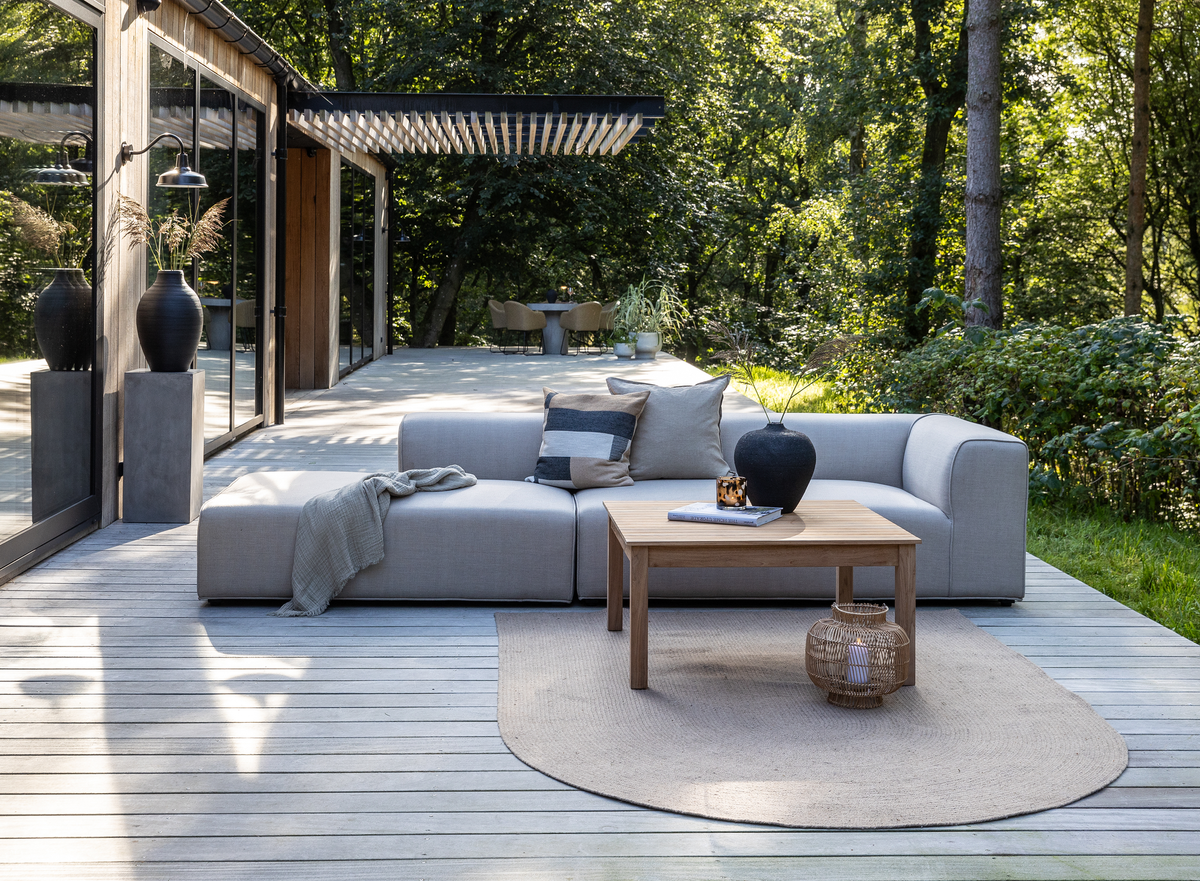 Moderne terrasse med grå modulsofa, træbord og lyst ovalt tæppe på trædæk, omgivet af grøn beplantning.