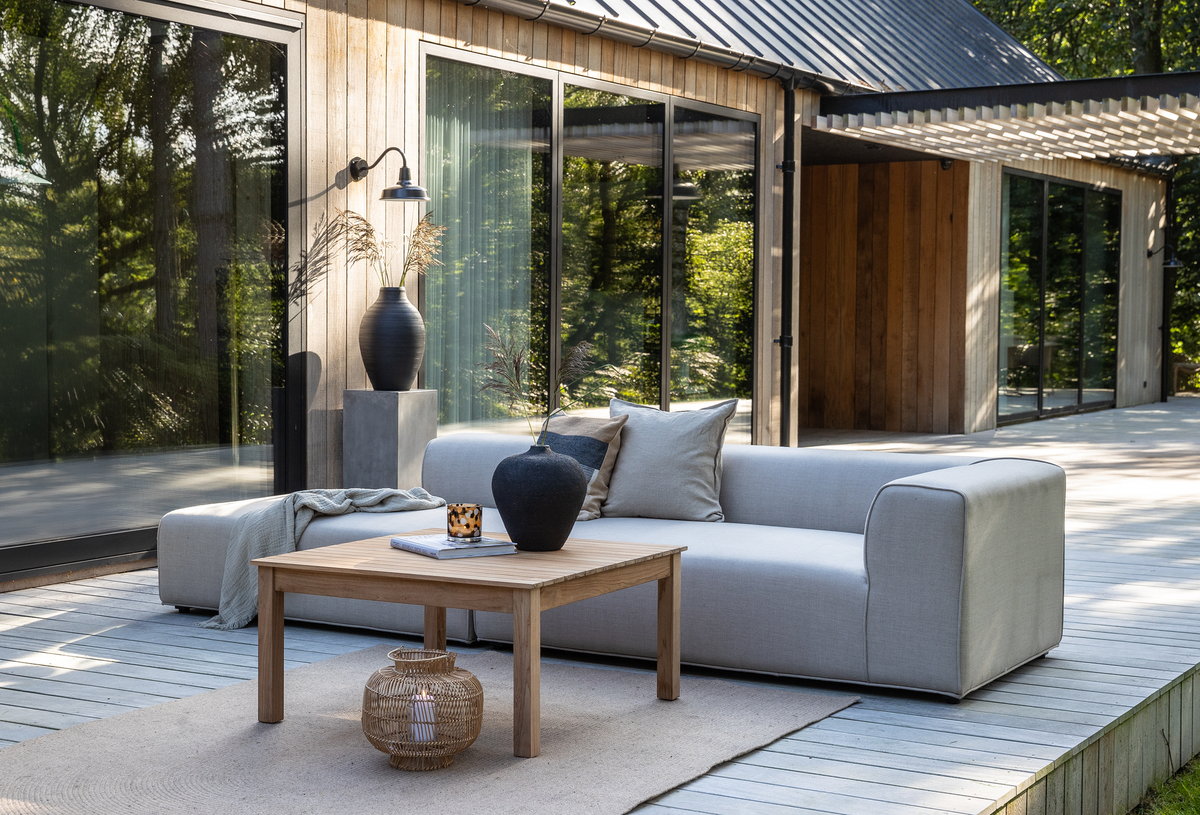 Moderne terrasse med lysegrå sofa, træbord og vaser foran et træhus med store glaspartier i dagslys.