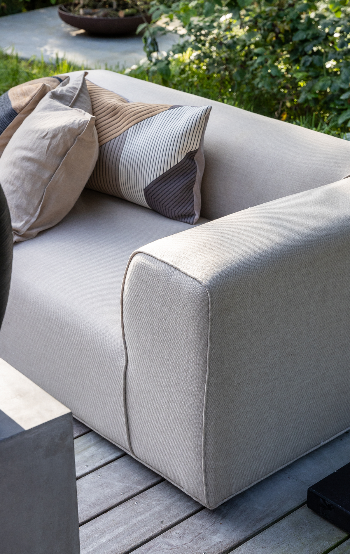 Lysebeige udendørs sofa med grafiske pyntepuder på en træterrasse i en grøn have.
