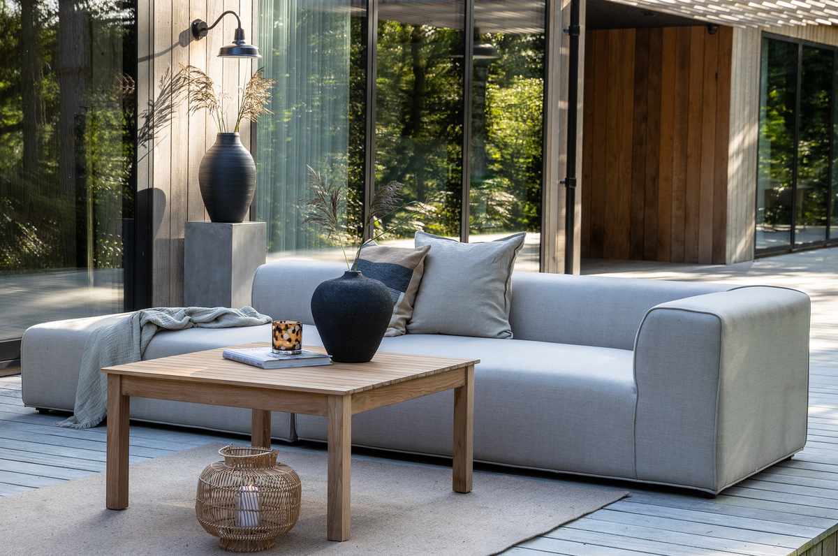Moderne terrasse med lysegrå sofa, træbord, sorte vaser og en kurvelygte på tæppe, ved et hus med glasvægge i dagslys.