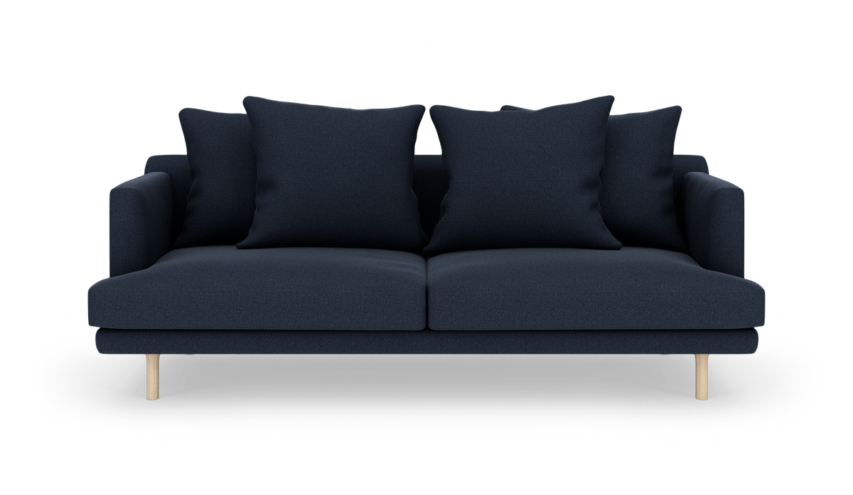 Mørkeblå sofa med fire store puder og lyse træben, set forfra mod hvid baggrund.