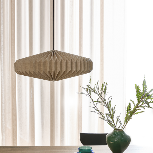 Foldet beige pendellampe over et bord med grøn vase og grene foran lyse gardiner.