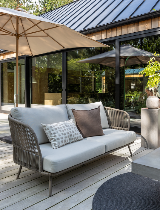 Udendørs sofa på træterrasse med puder under parasol ved glasdøre.
