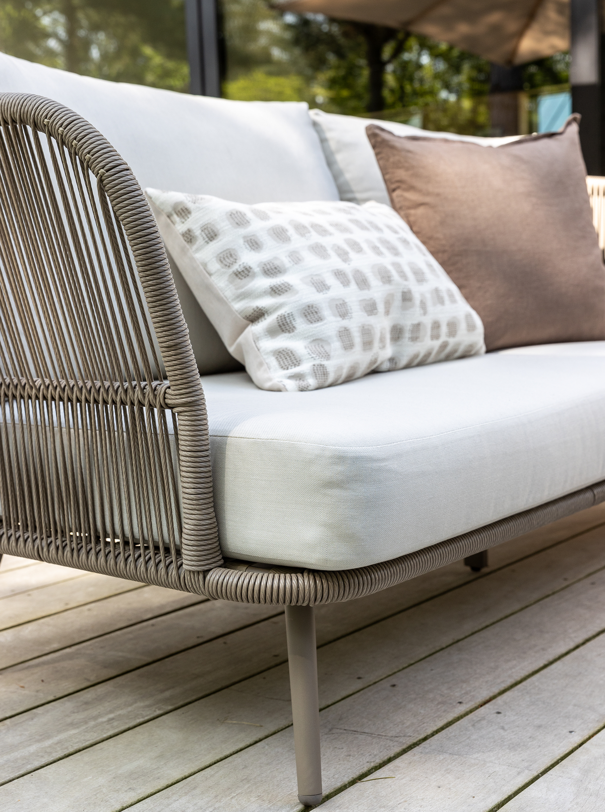 Nærbillede af moderne udendørs sofa i reb med lyse hynder og puder på en træterrasse.