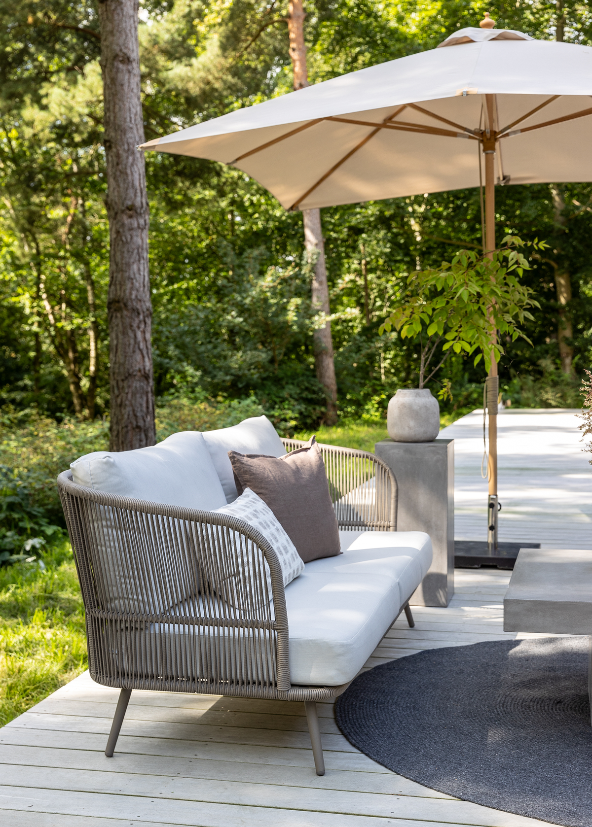 Lys terrasse i træ med flettet sofa med hvide hynder og puder, mørk rund måtte, betonbord og parasol omgivet af grøn natur.
