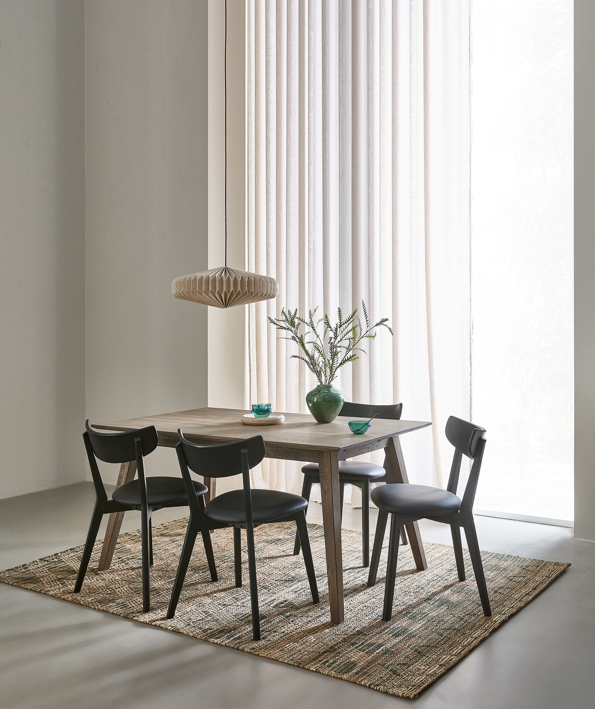 Spiseplads med træbord, fire sorte stole, vævet tæppe, grøn vase med grene og lyse gardiner i dagslys; beige pendel over bordet.