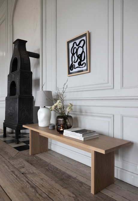 Lys skandinavisk indretning med en træbænk ved en panelvæg, bordlampe, vase med blomster, mørk glasvase og stakkede bøgerne ”This Is Home”; en sort støbejernsovn og et abstrakt vægbillede.