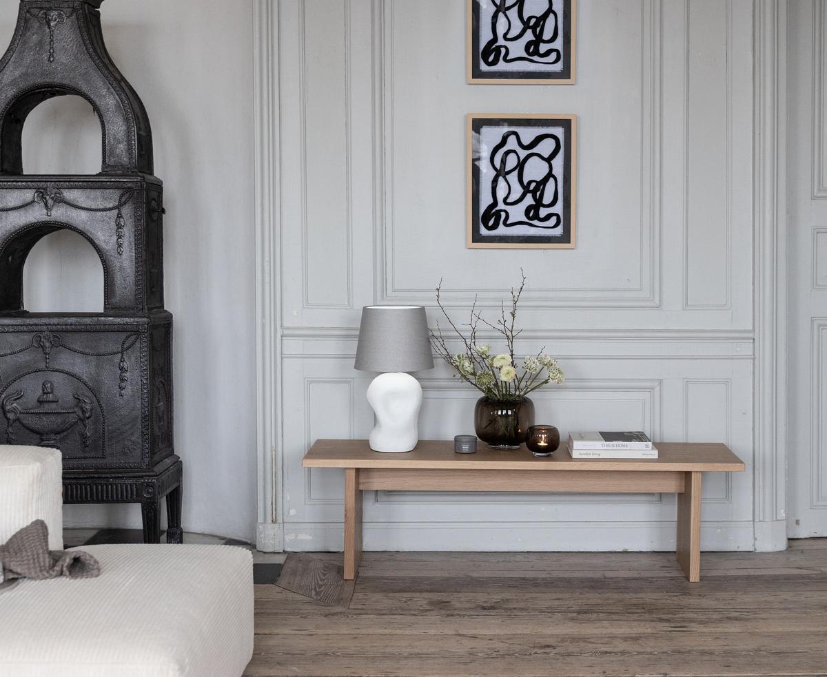 Lysegrå panelvæg med træbænk, bordlampe, glasvase med grene og blomster, lys og bøger; to abstrakte sort-hvide værker over samt en støbejernsovn og beige sofa i en rolig skandinavisk stue.