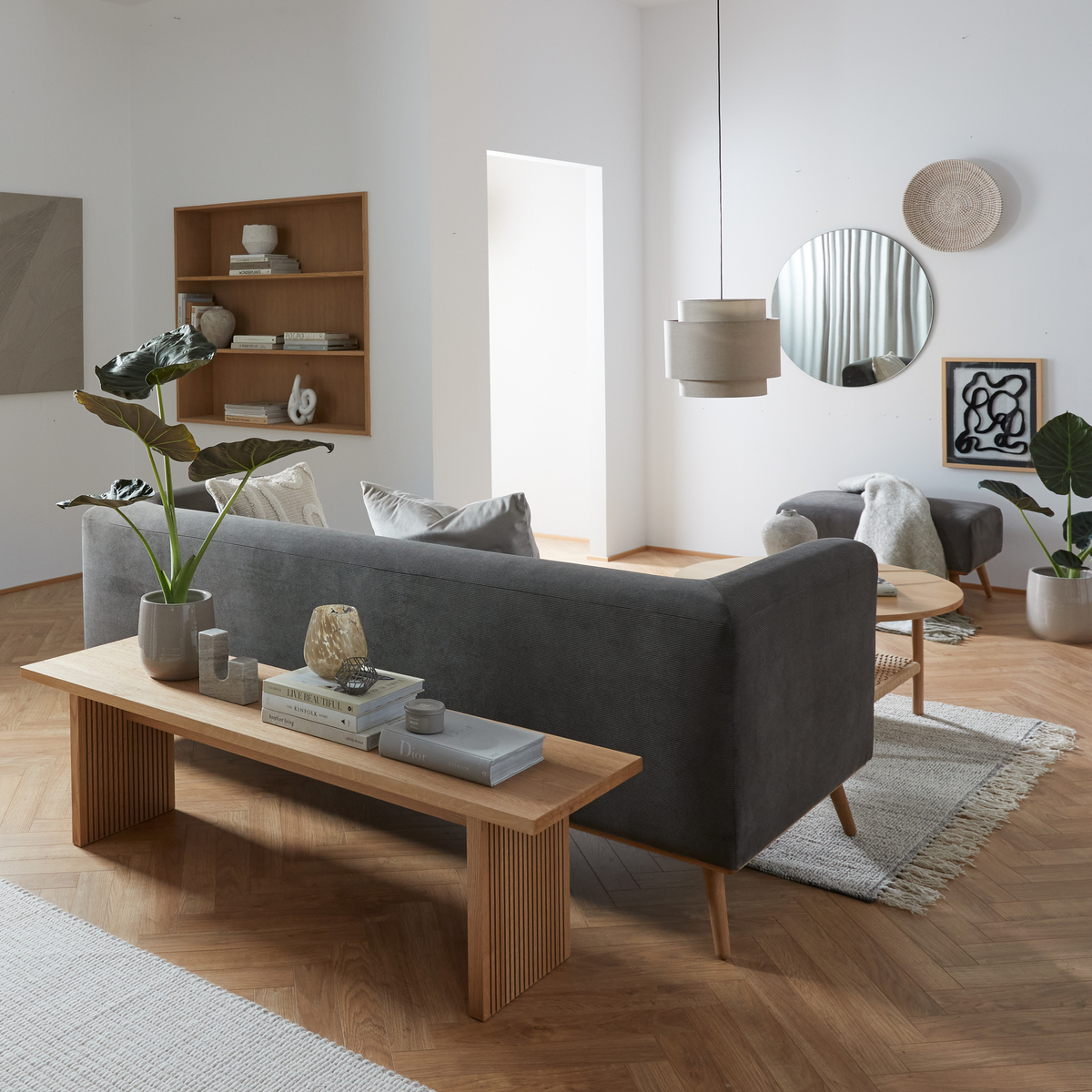 Lyst skandinavisk stue med grå sofa, bænk i lyst træ, rund spejl, pendellampe, planter og neutrale nuancer.