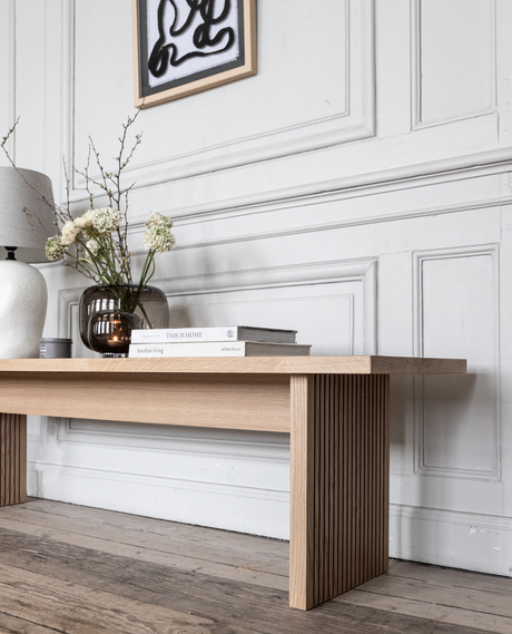 Lys træbænk foran hvid panelvæg med abstrakt kunst over; på bænken står en brun glasvase med hvide blomster, en hvid bordlampe og to bøger med titlerne ”THIS IS HOME” og ”barefoot living” – rolig, minimalistisk skandinavisk indretning.