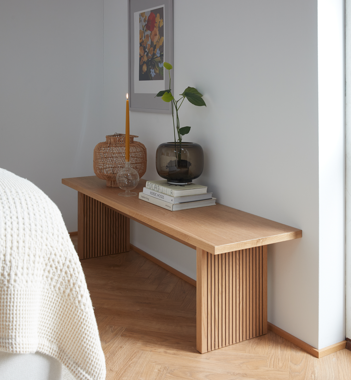Lyst hjørne i soveværelset med egetræsbænk pyntet med tændt slankt lys, glasvase med grøn plante, flettet lanterne og en stak bøger med titlerne “COLOR” og “NORDIC MOODS” under en indrammet blomsterplakat.