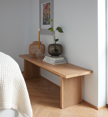 Lyst hjørne i soveværelset med egetræsbænk pyntet med tændt slankt lys, glasvase med grøn plante, flettet lanterne og en stak bøger med titlerne “COLOR” og “NORDIC MOODS” under en indrammet blomsterplakat.