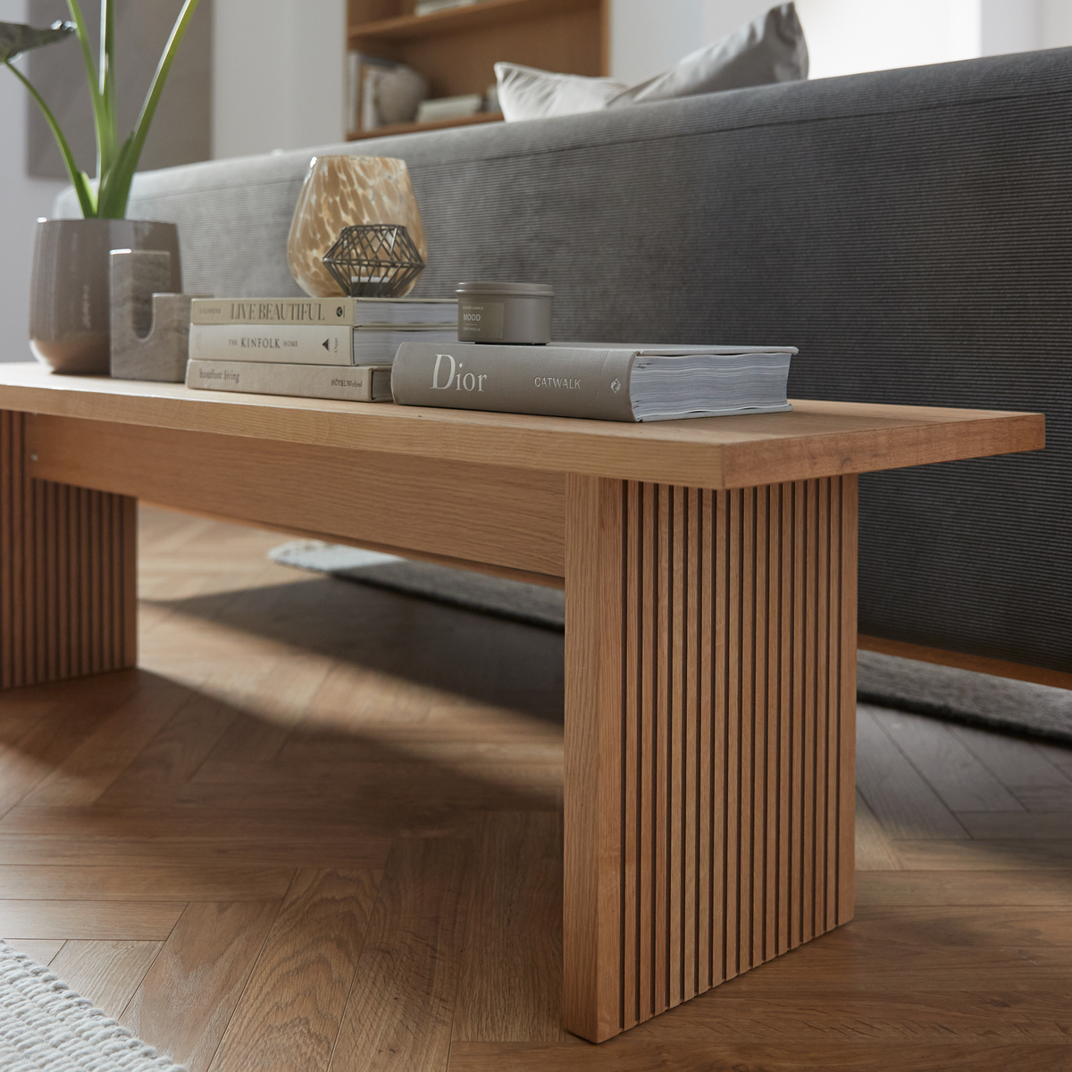 Lav bænk/sofabord i eg med lameller i en stue; på bordet ligger bøgerne Dior Catwalk, The Kinfolk Home og Live Beautiful samt lys, vase og en lille plante.