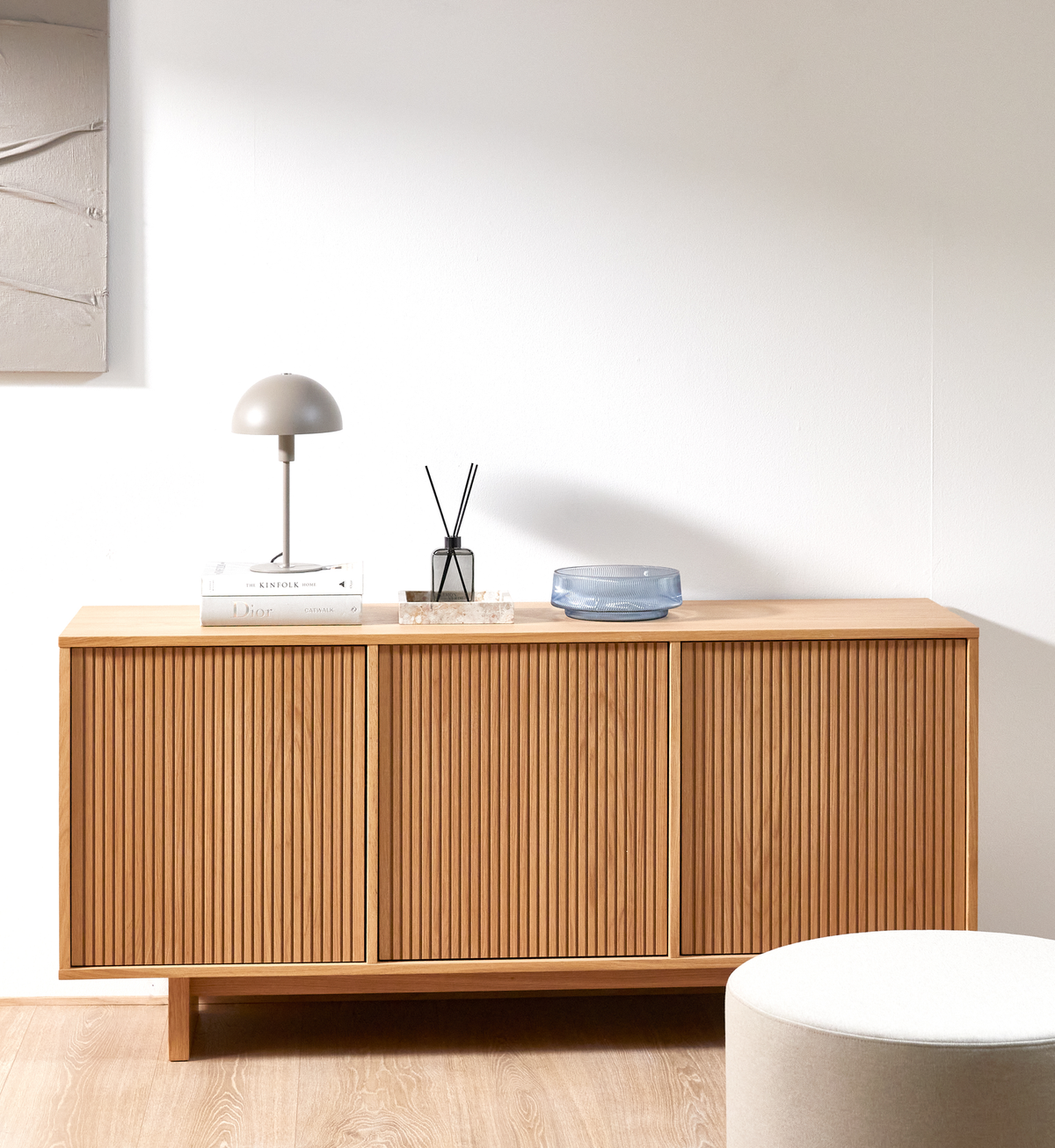 Minimalistisk stue med træskænk med lameller, bordlampe, Kinfolk- og Dior-bøger, duftpinde på stenbakke og blå glasskål mod en hvid væg.