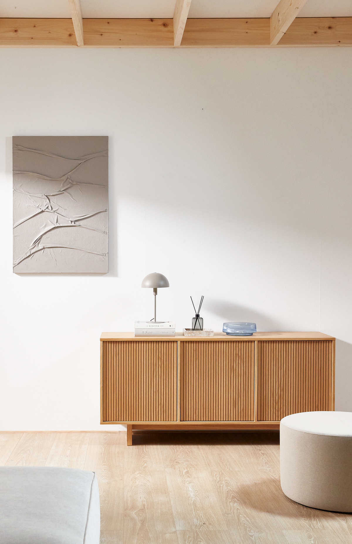 Lyst, minimalistisk stue i skandinavisk stil: ribbet træskænk med lille grå bordlampe på bøger med Dior, duftpinde og blå skål; abstrakt beige vægkunst og lyse trætoner.
