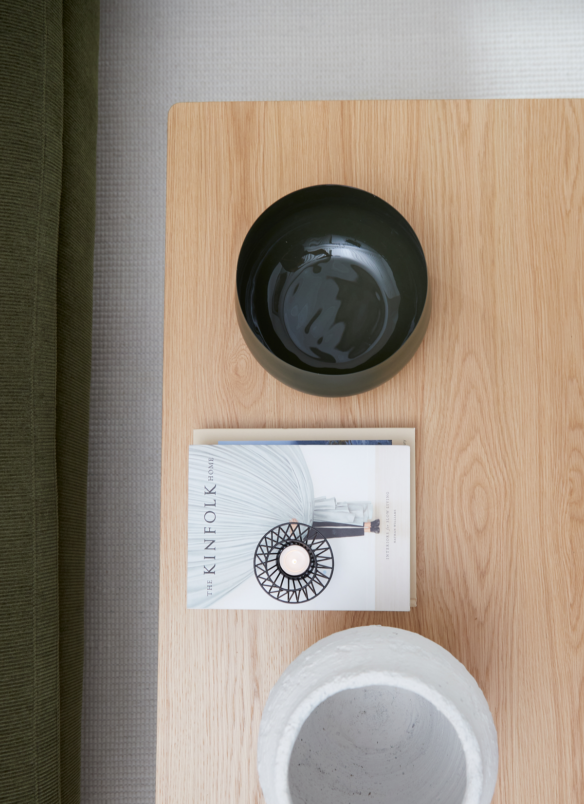 Lyst træbord med en mørk skål, et fyrfadslys i sort holder oven på bogen THE KINFOLK HOME og en lys betonvase; grøn sofa i kanten.