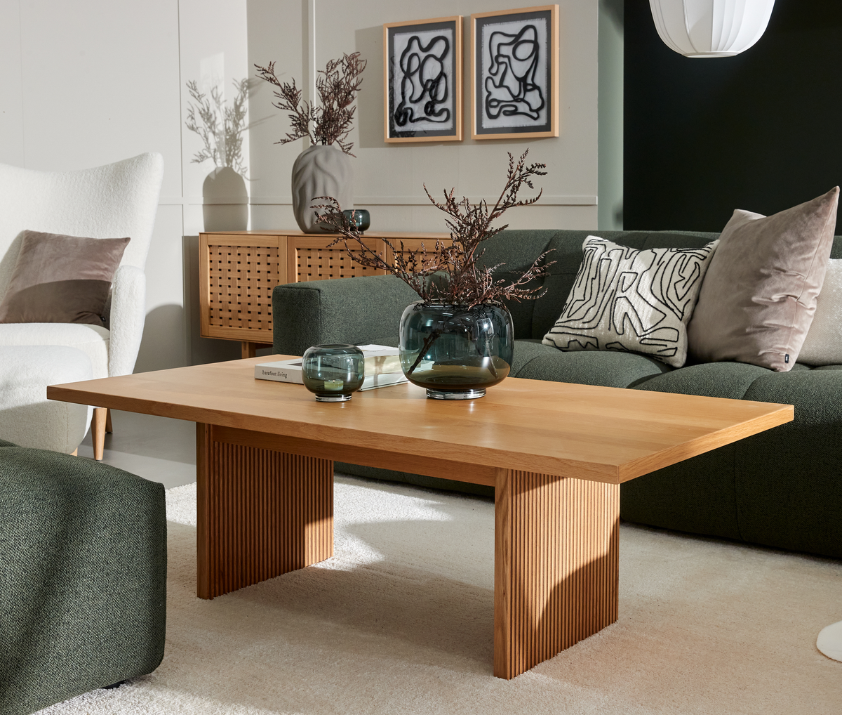Moderne stue: egetræsbord med riflede ben, grøn sofa med mønstrede puder, glasvaser med tørrede grene, to abstrakte billeder og en pendellampe i blødt dagslys.