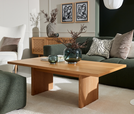 Moderne stue: egetræsbord med riflede ben, grøn sofa med mønstrede puder, glasvaser med tørrede grene, to abstrakte billeder og en pendellampe i blødt dagslys.