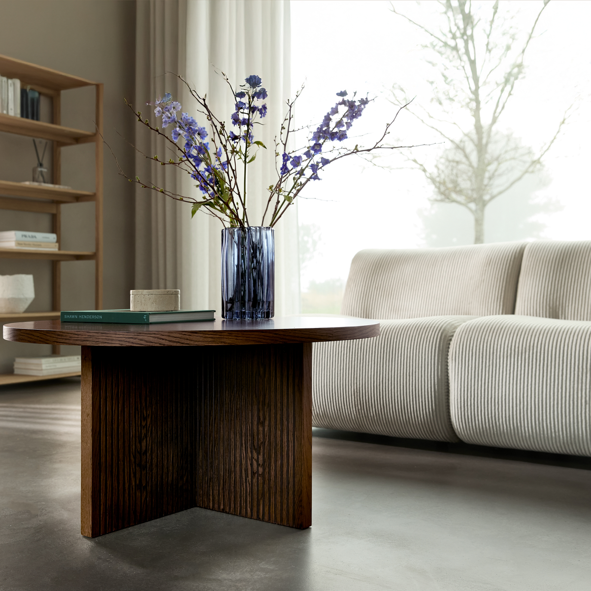Mørkt sofabord i træ med blå glasvase med lilla blomster og en bog foran en lys, rillet sofa ved et stort vindue med lette gardiner.
