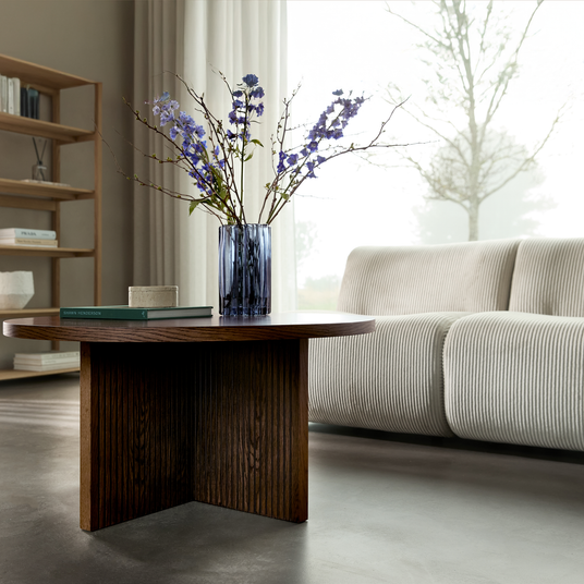 Mørkt sofabord i træ med blå glasvase med lilla blomster og en bog foran en lys, rillet sofa ved et stort vindue med lette gardiner.