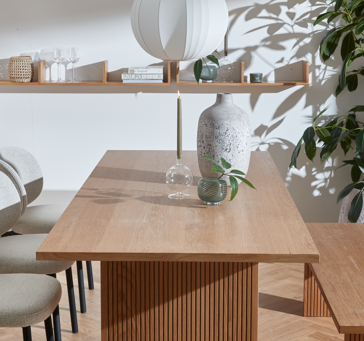 Lys spiseplads med egetræsbord med riflet sokkel, grå keramikvase, lille grøn vase med kvist og slank lys i glasstage; hylde med glas, bøger og dekorationer, hvid pendel og grønne planter i blødt dagslys.