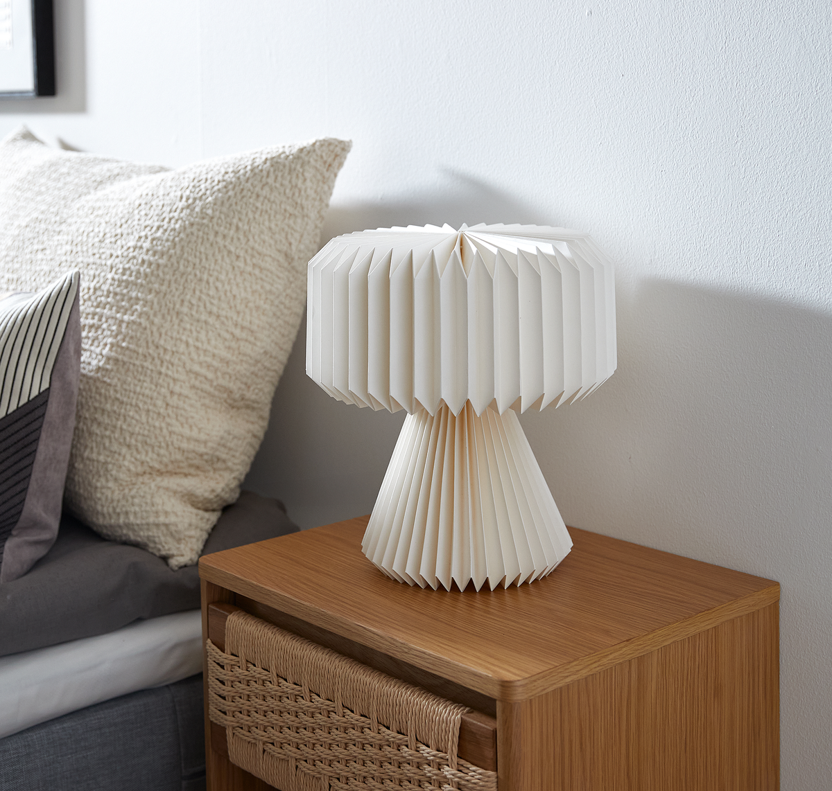 Hvid plisseret bordlampe i papir på natbord i eg med flettet skuffe ved siden af en seng med puder.