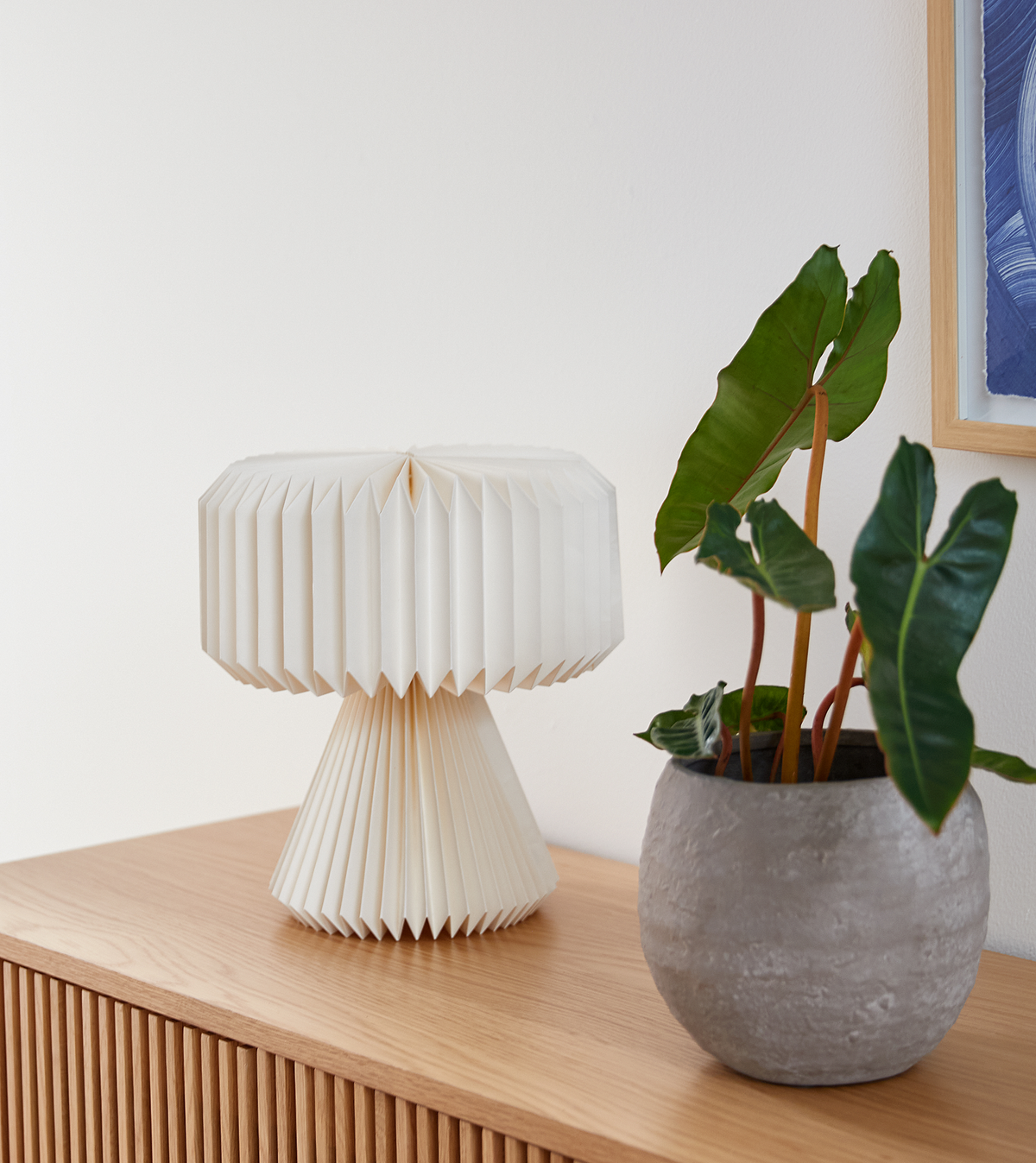 Hvid plisseret bordlampe og grøn potteplante på en rillet træskænk.