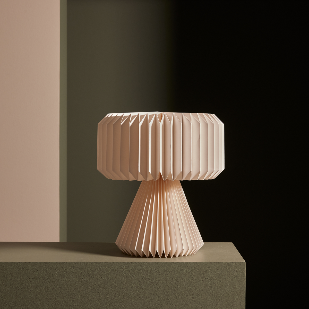 Plisseret bordlampe i lys rosa/beige på sokkel mod mørk, minimalistisk studie-baggrund.