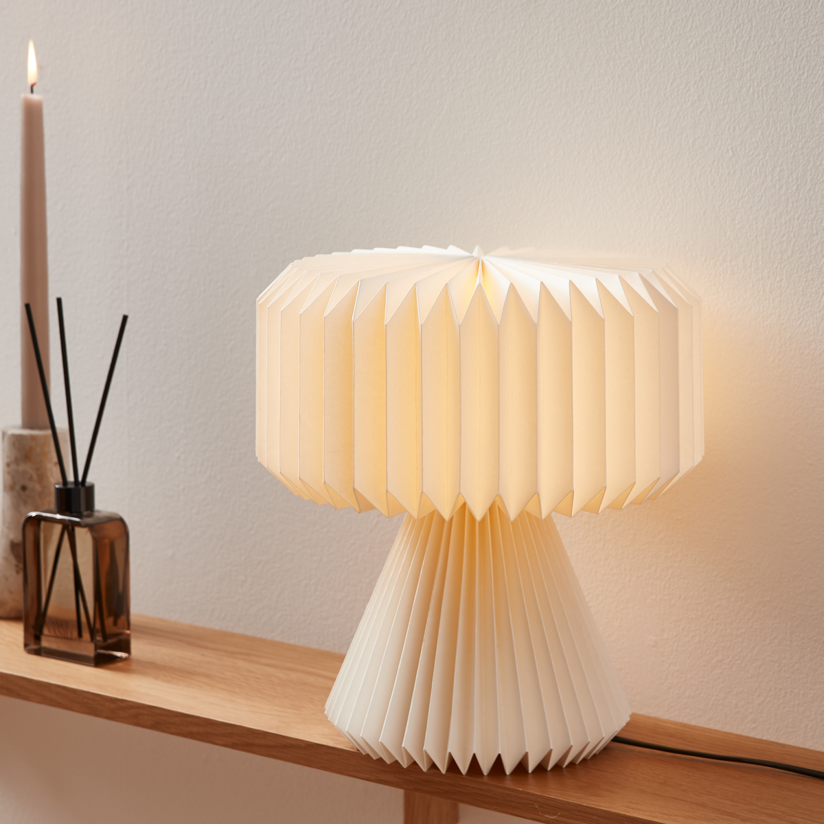 Foldet bordlampe i cremefarvet papir på en træhylde, tændt, ved siden af duftpinde og et smalt lys.