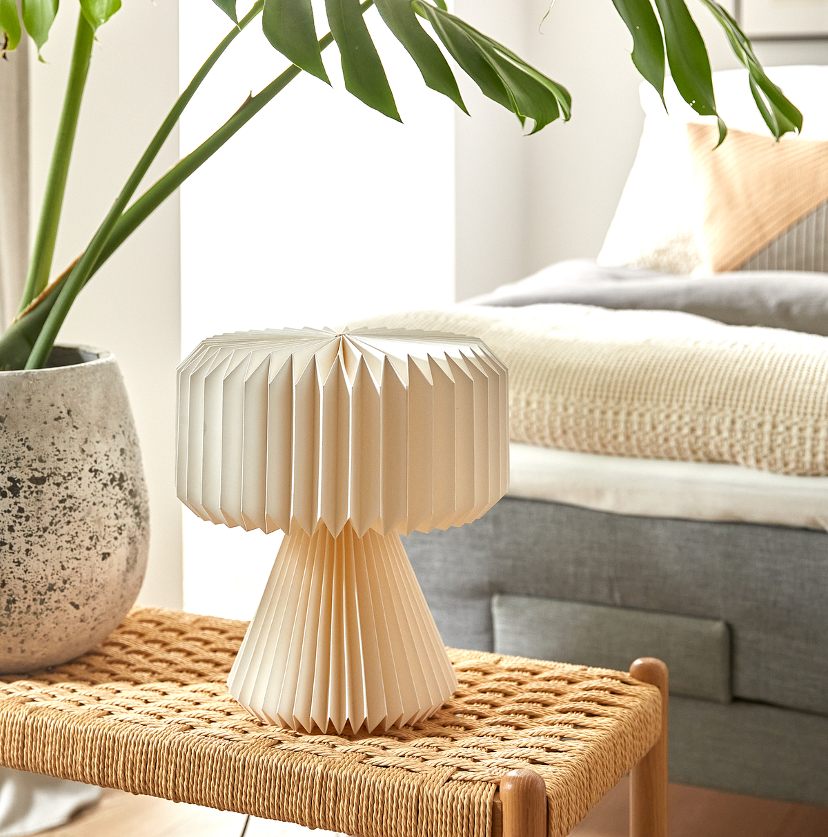 Cremefarvet, foldet bordlampe på en flettet bænk i et lyst soveværelse med strikket plaid, puder og en grøn potteplante.