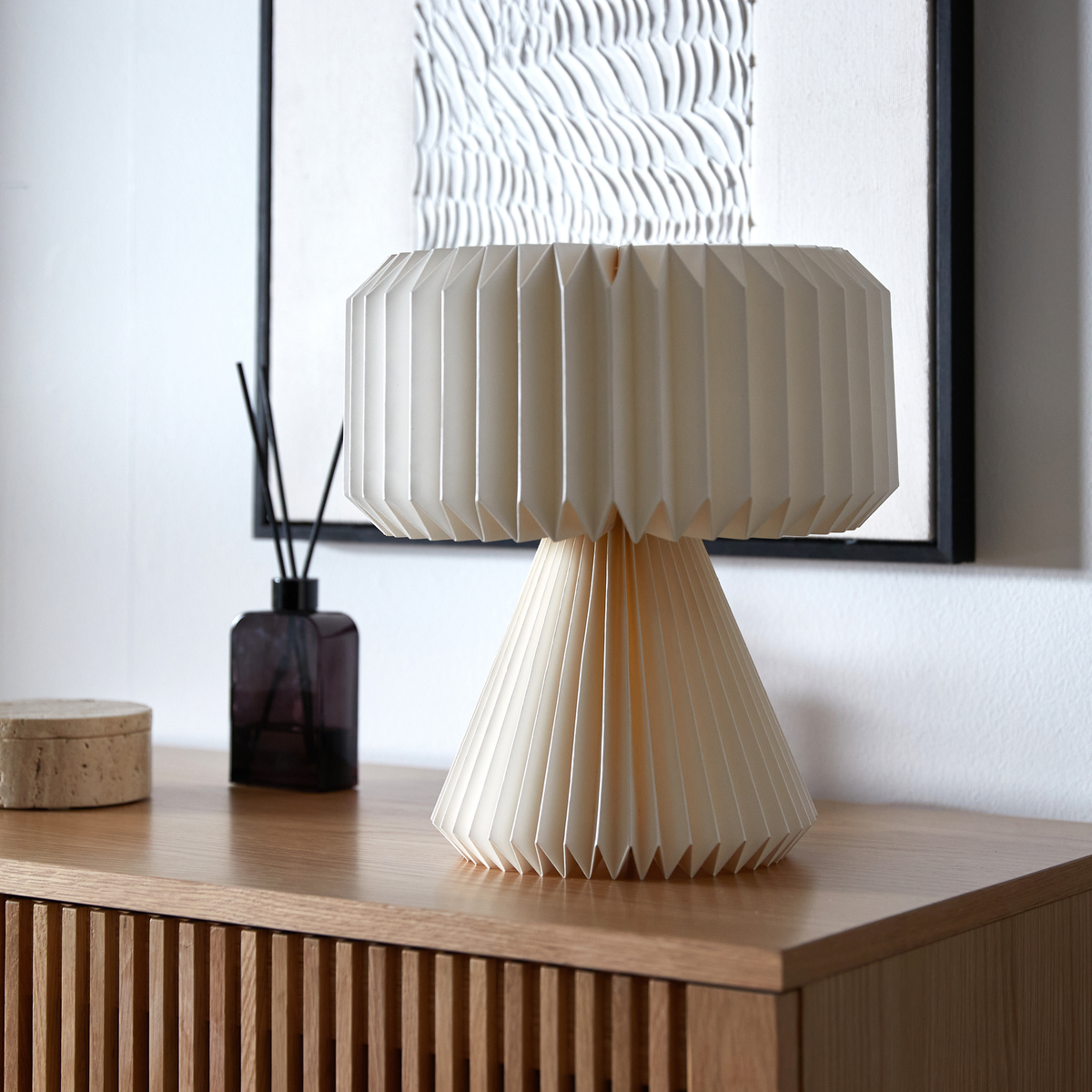 Foldet bordlampe i cremefarvet papir på egetræskommode, med sort duftpindeflaske og struktureret vægkunst i baggrunden.