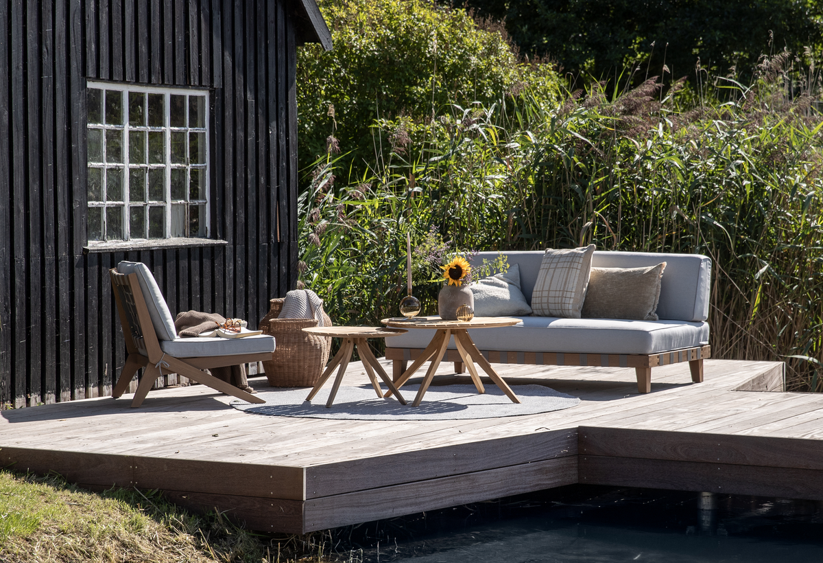 Udendørs trædæk ved vandet ved siden af et sort træhus; grå loungesofa og stol i træ med puder, to runde borde med vase med solsikke, lys og kurv med plaid i sollys.
