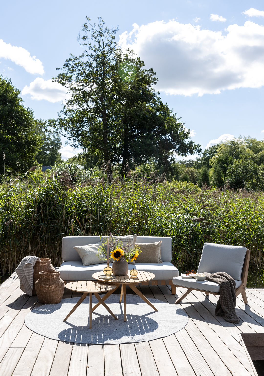 Lys terrasse på trædæk med grå sofa og loungestol, to runde sofaborde i træ med vase med solsikker, kurve, plaid og puder; sommerdag med blå himmel og skyer foran høje siv og træer.
