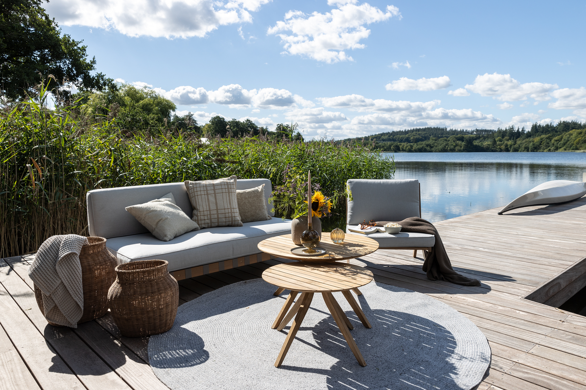 Udendørs terrasse ved sø med grå loungesofa, loungestol og runde træborde, kurve og rund måtte i solskin.