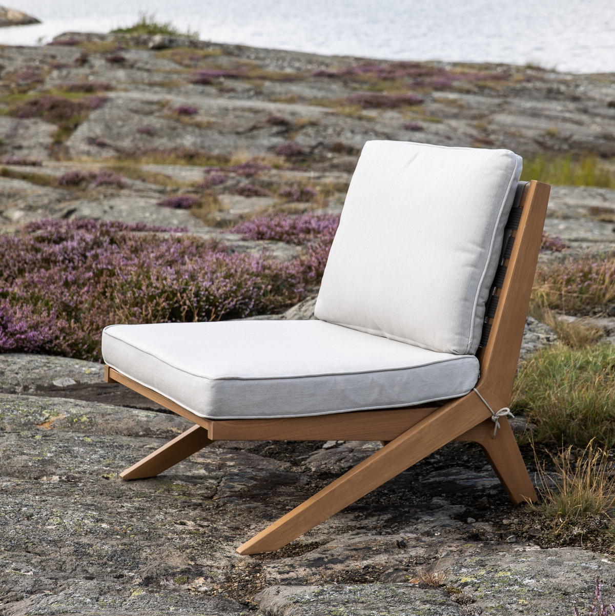 Lav loungestol i træ med lysegrå hynder placeret på klipper med lyng ved vandet i dagslys.