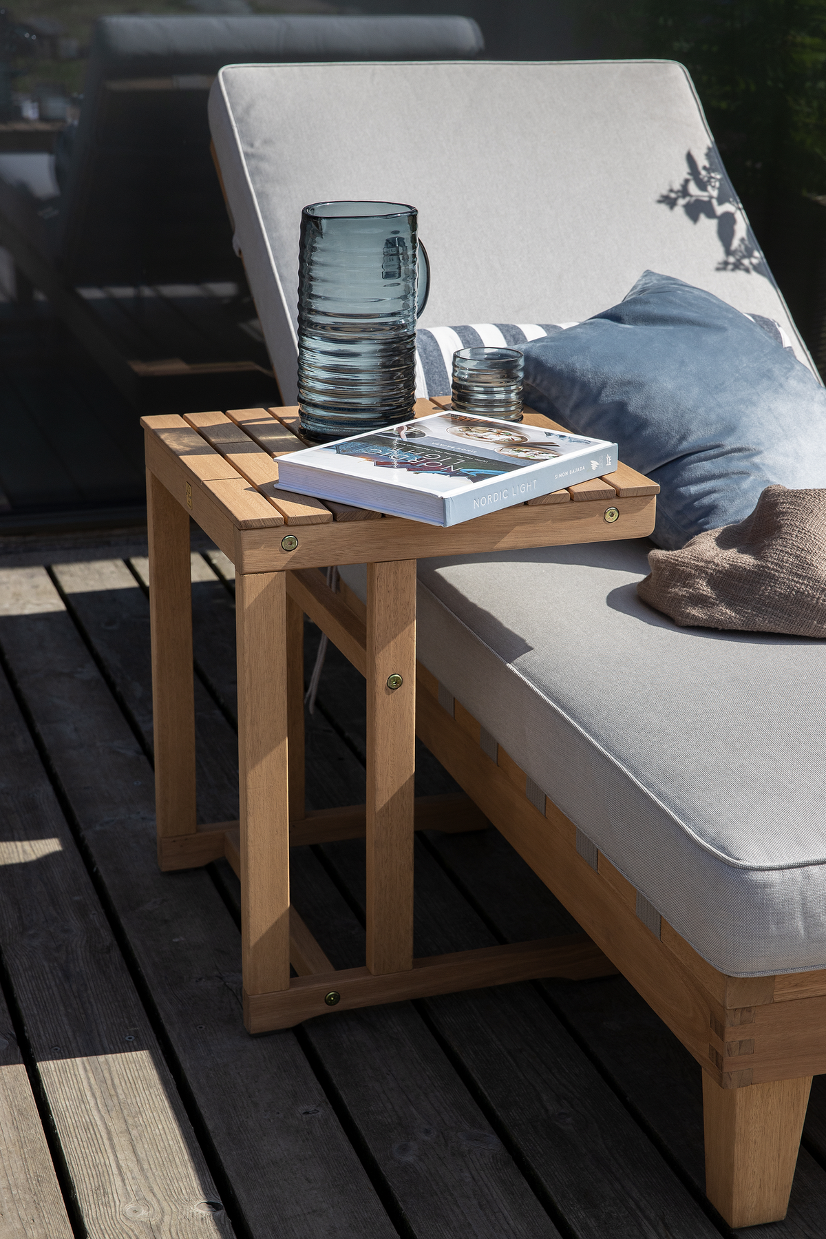 Træsidebord ved en grå solseng på terrasse med blå pude; en riflet glas karaffel, et glas og en bog med titlen Nordic Light ligger i solen.
