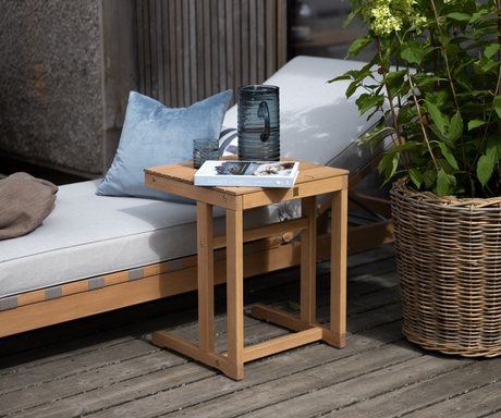 Udendørs miljø med solseng og lille træbord på terrasse; på bordet står en blå glas-kande, et glas og bogen ”Nordic Light”, ved siden af en fletkurv med grøn plante.