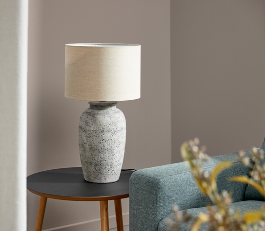 Bordlampe med beige stofskærm og stenlignende fod på rundt sidebord ved siden af turkis sofa.
