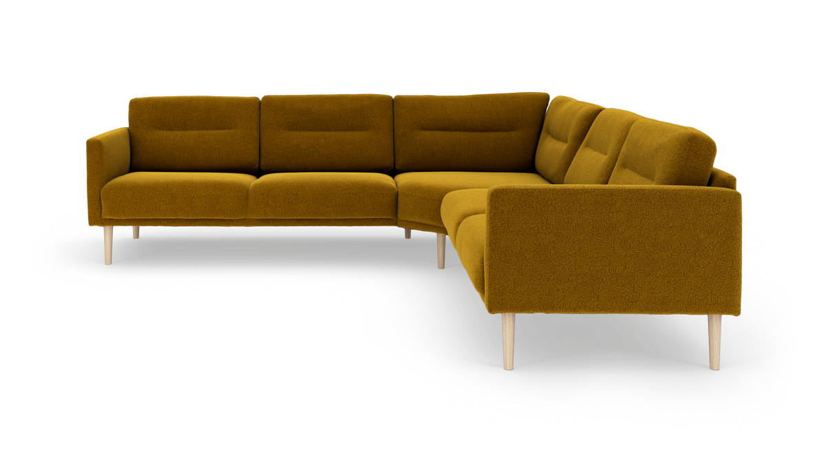 Modulær L‑sofa i sennepsgul tekstil med lyse træben, vist i studie fra skråt forfra.