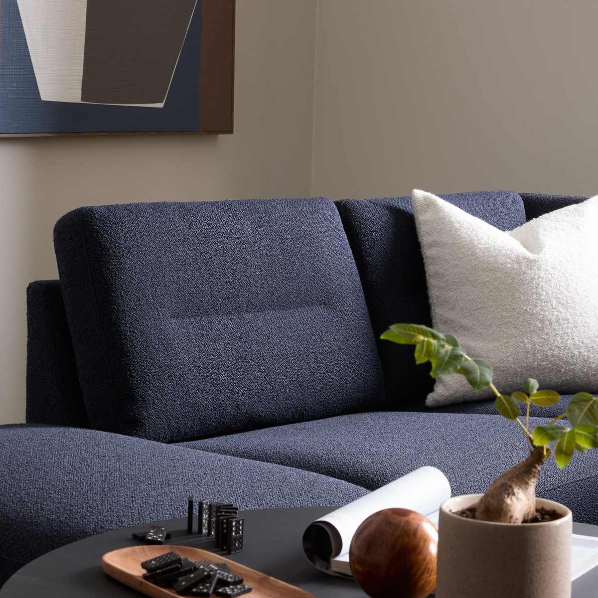 Mørkeblå sofa med hvid tekstureret pude, sort sofabord med bakke og dominobrikker, lille potteplante og vægkunst i baggrunden.