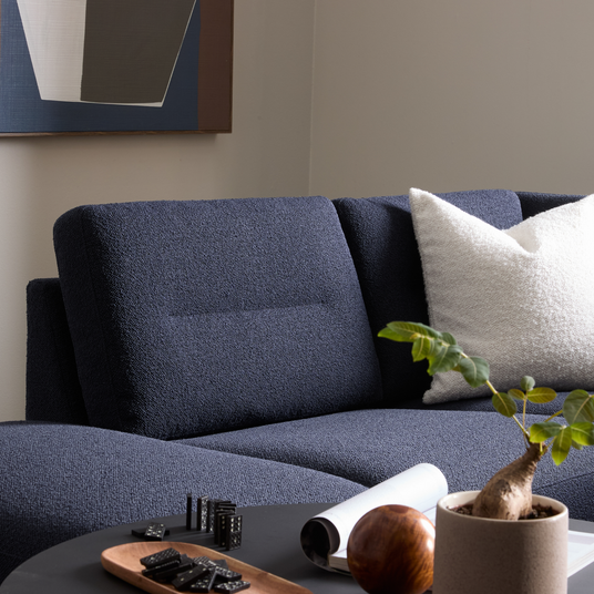 Mørkeblå sofa med hvid tekstureret pude, sort sofabord med bakke og dominobrikker, lille potteplante og vægkunst i baggrunden.