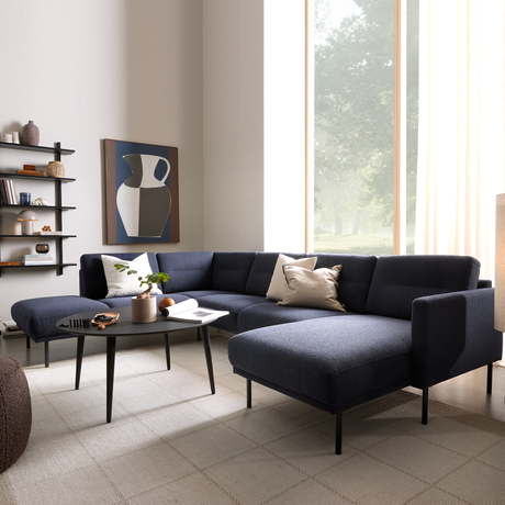 Moderne stue med mørkeblå hjørnesofa med chaiselong, sort sofabord med plante, væghylder og abstrakt vasebillede ved stort vindue med lyse gardiner i blødt dagslys.