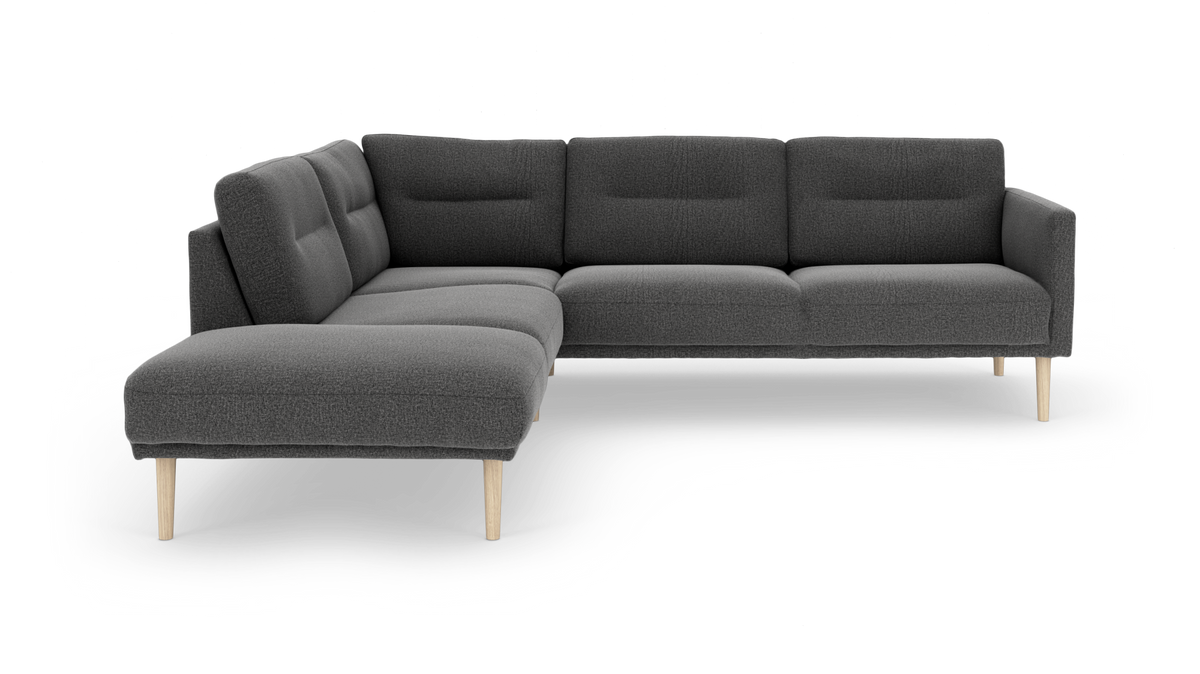 Mørkegrå L‑sofa med chaiselong og lyse træben, i studiemiljø.