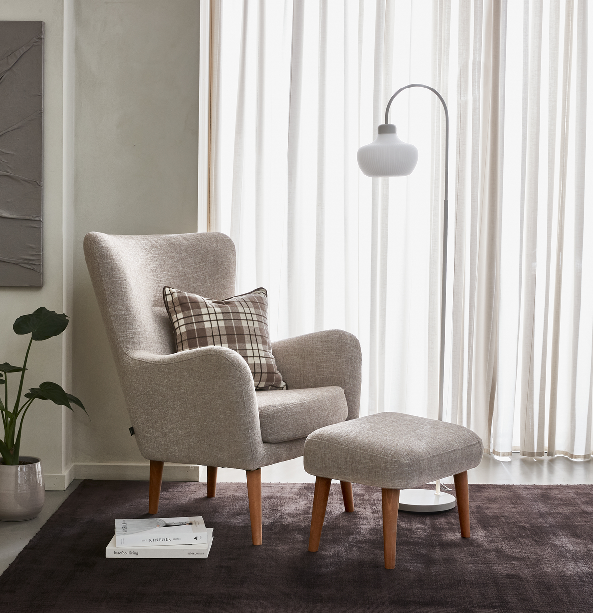 Lænestol i beige stof med skammel, ternet pude, gulvlampe og bøgerne The Kinfolk og Barefoot Living på mørkt tæppe ved lyse gardiner.