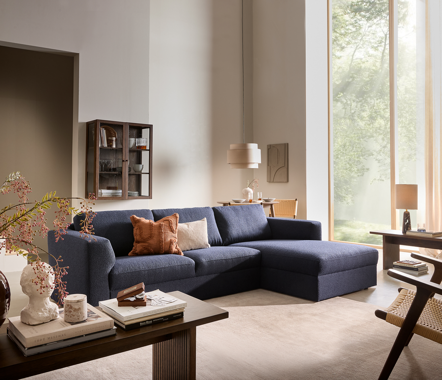 Lyst stue med en mørkeblå hjørnesofa, rust- og beige puder, beige tæppe, træborde med bøger og vase, pendel og store vinduer med lette gardiner i dagslys.