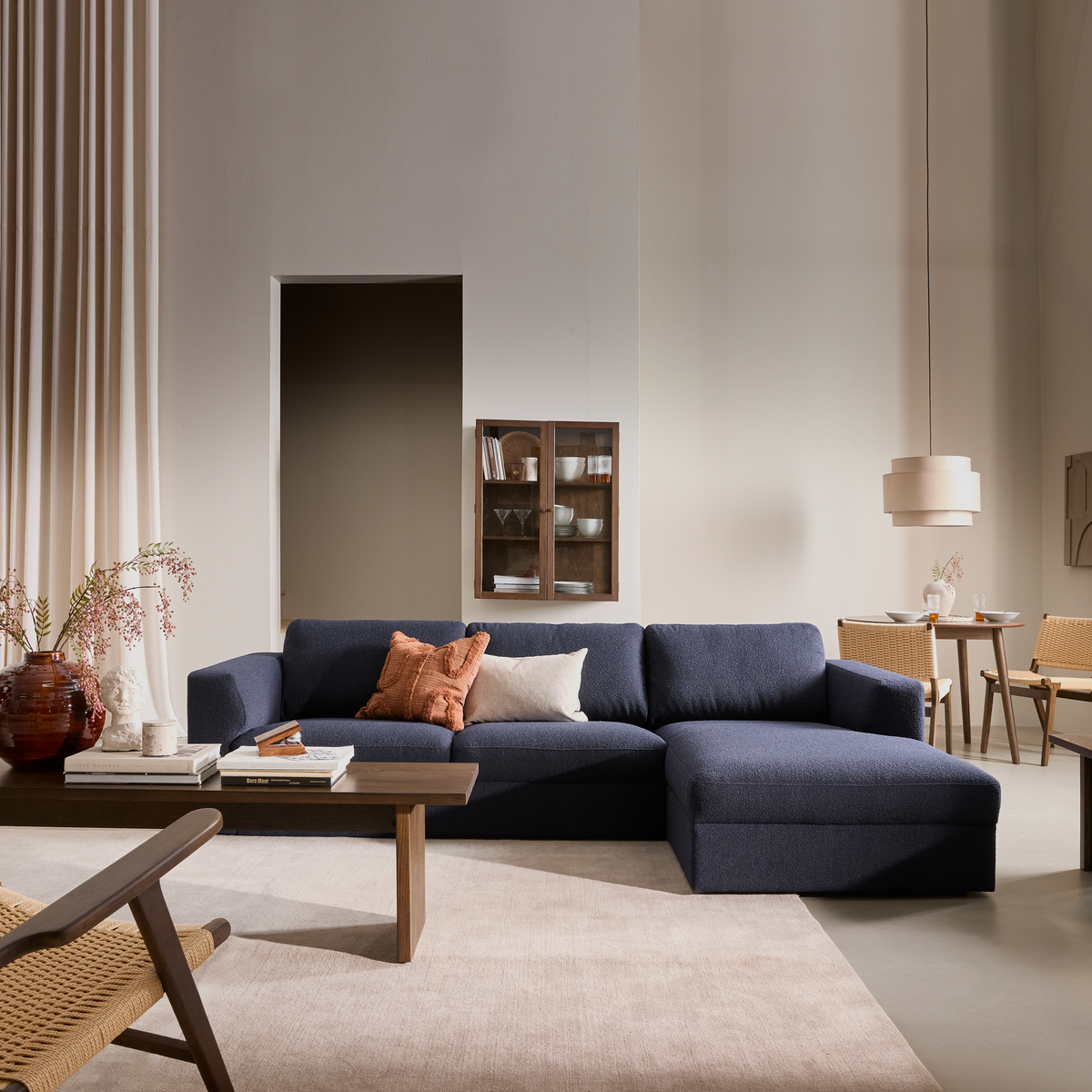 Beige stue i blødt dagslys med mørkeblå hjørnesofa, træsofabor, puder, tæppe, gardiner, vægskab og en lille spiseplads med fletstole og beige pendel.